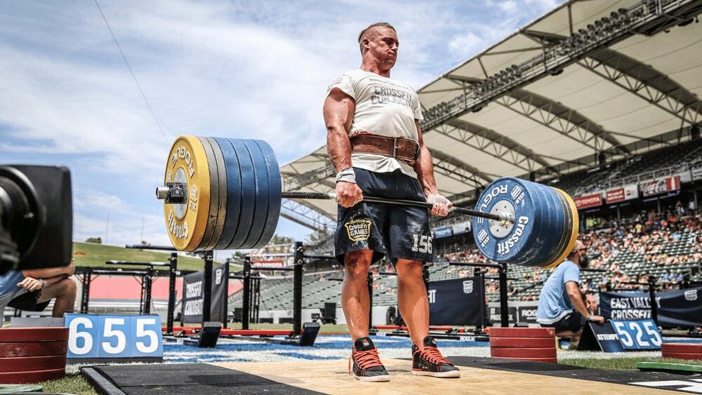 Man performing a 655-pound deadlift at a CrossFit competition in an outdoor stadium, using a Rogue barbell with bumper plates.