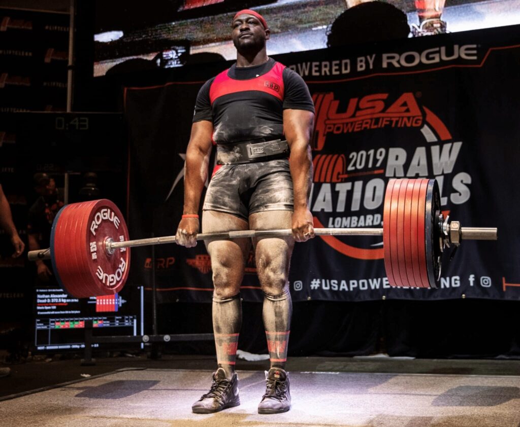 Muscular Black man in a red and black outfit deadlifting a heavy barbell on a powerlifting stage.