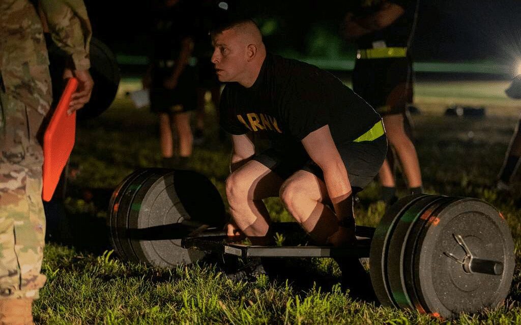 Man in a black "ARMY" t-shirt and shorts performing a trap bar deadlift outdoors at night.
